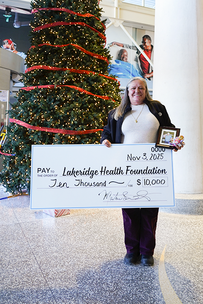 Martine holding a photo of Danny and a cheque for Lakeridge Health Foundation, standing in front of a Christmas tree within the Hospital.